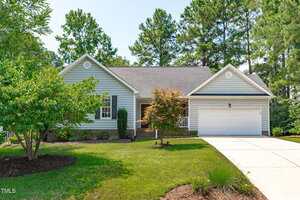 A single-story house with gray siding, a covered front porch, and a two-car garage. The front yard has green grass, shrubs, and small trees, with a concrete driveway leading to the garage.