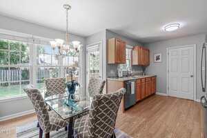 A kitchen and dining area with light wooden floors, a glass dining table with four patterned chairs, wooden cabinets, stainless steel appliances, large windows, and a door leading outside.