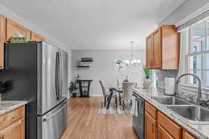 A modern kitchen with stainless steel appliances, light wood cabinets, granite countertops, and a double sink. The space opens to a dining area with a table, chairs, wall art, and a window letting in natural light.