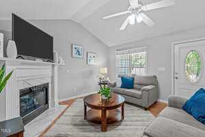 A living room with light gray walls, a ceiling fan, a fireplace with a TV above it, two gray armchairs with blue pillows, a round wooden coffee table, and a door with an oval window.