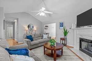 A living room with two gray sofas, a round wooden coffee table, green plants, a fireplace with decor, wall art, a ceiling fan, and a view into a hallway and another room. The room has light walls and wood flooring.