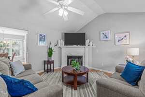 A living room with gray walls, a ceiling fan, a fireplace with a TV on top, two gray sofas with blue pillows, a round wooden coffee table, a rug, framed artwork, and a floor lamp.