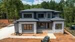 A two-story modern house under construction at 3921 Cedar Knolls Drive in Youngsville features gray siding, stone accents, large windows, a covered front porch, and driveway. Construction materials and equipment are visible on site with trees in the background.