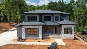 A two-story modern house under construction at 3921 Cedar Knolls Drive in Youngsville features gray siding, stone accents, large windows, a covered front porch, and driveway. Construction materials and equipment are visible on site with trees in the background.