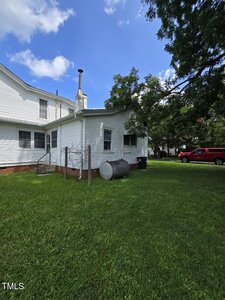 A white two-story house with a small attached section, metal fuel tank, and green lawn. A red vehicle is parked near trees under a blue sky with scattered clouds.