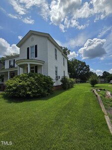 Two-story white house with black shutters and a small front porch, surrounded by green grass and shrubs, under a partly cloudy sky. A wooden beam borders part of the lawn on the right side.