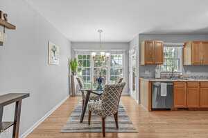 A dining area with a glass table and four patterned chairs on a striped rug, next to a kitchen with wooden cabinets and granite countertops. Large windows let in natural light, and a plant sits by the window.
