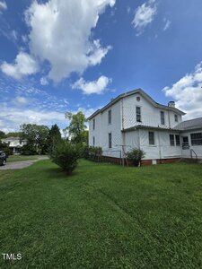 A two-story white house with a gabled roof sits on a grassy lawn under a partly cloudy sky. There are some trees and a driveway in the background. The house has several windows and a brick foundation.