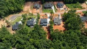 Aerial view of several houses in a residential neighborhood with some homes under construction, surrounded by trees and patches of red soil. A road runs horizontally through the center of the image.