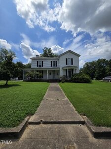 A two-story white house with black shutters is centered behind a concrete walkway, surrounded by green grass, shrubs, and trees, under a partly cloudy blue sky.