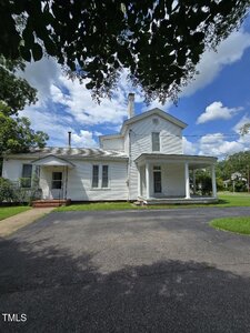A two-story white house with a covered front porch and a chimney is shown under a sunny sky with scattered clouds. The house has white siding, several windows, and sits next to a driveway and lawn with trees.