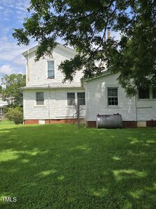 A white two-story house with an attached single-story section, seen from the side, with a large metal tank next to it and a green lawn in the foreground. Trees and blue sky with clouds are visible.