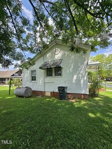 A white house with a metal awning over one window, a black trash bin, and a large metal tank sits on a grassy lawn with trees providing shade. The sky is partly cloudy and other houses are visible in the background.