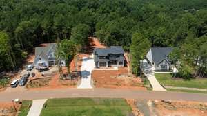 Aerial view of three newly built houses with driveways on a dirt lot, surrounded by trees and bordered by a paved road in a suburban neighborhood.