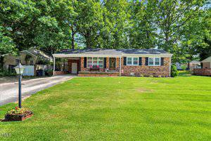 Charming single-story brick house at 549 Sagefield Drive in Henderson, featuring black shutters, a covered carport, and a cozy front porch. Enjoy the large lawn with a lamppost and flower bed, surrounded by mature trees.