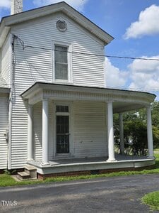 A white two-story house with vertical siding, a circular attic vent, and a covered porch supported by four columns. There are two steps leading to the porch and a paved driveway in the foreground.