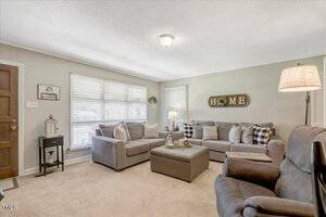 A cozy living room with two gray sofas, a gray recliner, an ottoman, and a floor lamp. Large windows with blinds let in natural light. Decorative pillows and a "HOME" wall sign add accents.