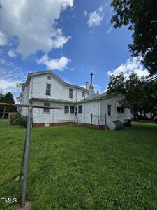 A white two-story house with a shingled exterior is shown from the backyard. The yard has green grass, a metal clothesline pole, an oil tank, and a tree on the right side under a partly cloudy sky.