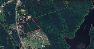 Aerial map view showing a outlined property bordered in red adjacent to a wooded area and body of water; surrounding lots, roads, and a trail marked with a dashed yellow line are also visible.