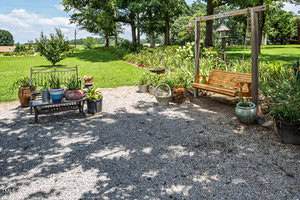 A gravel patio area with a black metal bench and cushions, several potted plants, a wooden swing on a frame, and a variety of garden decorations, surrounded by grass and trees on a sunny day.