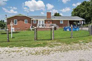 A single-story brick house with a fenced backyard, small wooden deck, above-ground pool, birdhouse, and green lawn under a partly cloudy sky. Gravel driveway in the foreground.