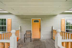 A front porch with a light gray siding exterior, a central orange front door with a small window, two windows with orange shutters, a single brown chair, and white railings on either side.