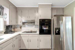 A kitchen with light gray cabinets, a stainless steel refrigerator, a black built-in double oven, a stovetop, a vent hood, and a brick backsplash. A coffee maker and toaster are on the counter.