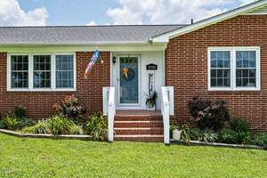 A brick house with white trim features a central glass front door with a "Welcome" sign, a small porch with steps, potted plants, and a rainbow pride flag by the windows. The yard is green with shrubs and flowers.