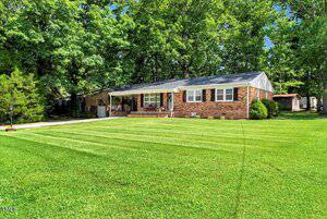 A single-story brick house with a covered front porch, white trim, and a large, well-maintained green lawn. The house is surrounded by tall trees and has a driveway on the left side.