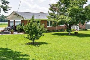A one-story brick house with a covered carport sits on a large, well-maintained lawn. Trees and shrubs are in the yard, and a patio area with chairs is visible near the carport. The sky is mostly clear.