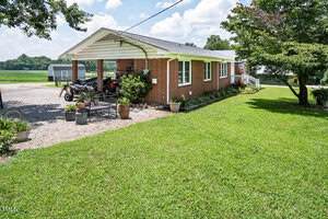 A single-story red brick house with a covered carport, various potted plants, and outdoor furniture sits on a well-maintained lawn with gravel driveway and an outbuilding in the background.