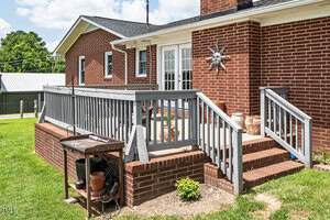 A red brick house with a gray wooden porch and steps, a decorative sun face on the wall, and potted plants and garden tools arranged near the railing. The yard is grassy with a few small plants.