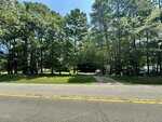 View of a paved road with double yellow lines in Henderson, bordered by grass and dense trees. A gravel driveway leads into the wooded 0 Tom Hicks Land, as sunlight filters through the branches.