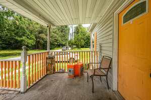 A covered porch with a yellow front door, a brown chair, orange coolers, and a wooden railing. The porch overlooks a grassy yard with trees and a parked car in the background.