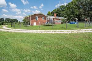 A one-story brick house with a chimney sits on a grassy lot enclosed by a wire fence. A gravel driveway curves around the yard, and a large tree provides shade near the house. A blue above-ground pool is visible in the yard.