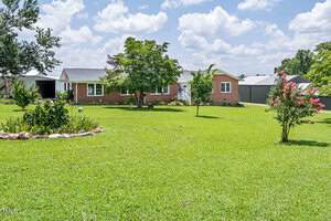 A one-story brick house with a gray roof is set back on a large green lawn. There are several small trees and landscaped areas in the yard, and metal buildings are visible in the background under a partly cloudy sky.