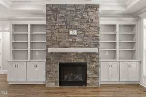 A stone fireplace with a white mantel is centered between two built-in white shelving units with cabinets below. The room has hardwood floors and white trim.