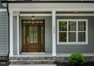 A modern house entry with a wooden front door featuring glass panels, gray horizontal siding, white trim, stone accents at the base, and the number 1224 vertically displayed beside a double window.