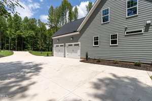 The image shows a large concrete driveway leading to a three-car garage attached to a gray house with white trim, surrounded by trees and landscaped with small shrubs.