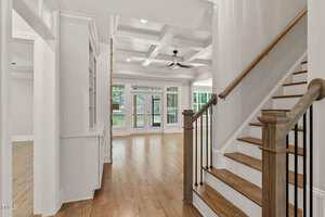 A view of a modern home interior featuring a wooden staircase with white risers and black balusters, coffered ceilings, hardwood floors, built-in shelves, and large windows overlooking a backyard.
