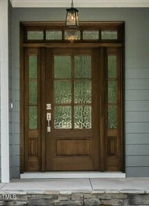 A brown wooden front door with decorative glass panels, a silver handle, and a matching lock, framed by gray siding and stone steps, with a lantern-style light fixture above.