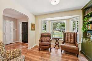 A living room with hardwood floors, two brown leather armchairs near a bay window, a small round table, a floral upholstered chair, green built-in shelves with books and plants, and an arched entryway leading to a foyer.