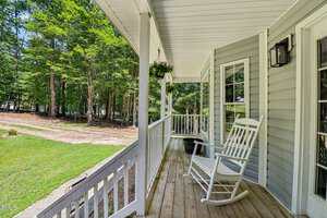 A covered wooden front porch with white railings features a white rocking chair and a hanging plant. The porch overlooks a grassy yard with trees in the background.
