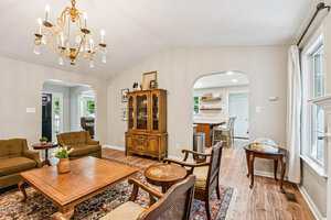 A living room with two armchairs, a wooden coffee table, and a vintage hutch. A globe sits near a window, and a chandelier hangs from the ceiling. The room opens into a kitchen with barstools and white cabinetry.