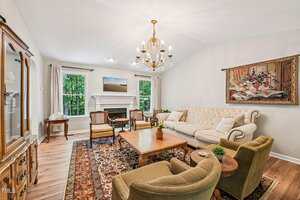 A living room with a beige sofa, two green armchairs, a wooden coffee table, and a patterned rug. There is a fireplace between two windows, a chandelier, and a framed painting on the wall.