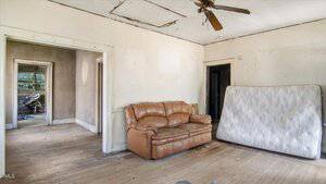 A worn brown sofa and a mattress rest against the walls of a sparsely furnished, rundown room with a damaged ceiling, wooden floor, and peeling paint. Open doorways lead to other similarly neglected rooms.