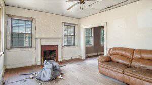 A worn living room with peeling paint, damaged ceiling, debris on the floor, an old fireplace, two windows, and a brown leather couch against the right wall. A torn chair is positioned near the fireplace.