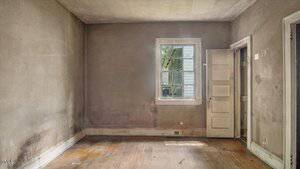 An empty, worn room with dirty walls, stained wooden floors, and a single window letting in light. The door is open, revealing another room. The space appears neglected and in need of repairs.