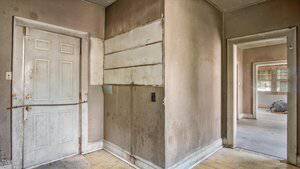 A dirty, partially damaged entryway with stained walls, a white door secured by a wooden plank, exposed wall sections, and an adjacent hallway leading to another empty, run-down room.