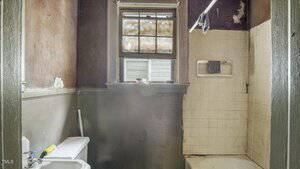 A small, worn bathroom with peeling paint, a dirty window, an old sink and toilet, and a tiled bathtub with visible grime and damage. A cleaning tool rests on the sink.